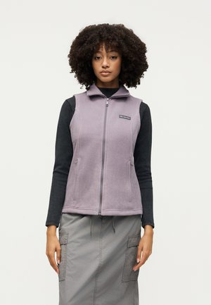 Γυναίκα με κυματιστά μαλλιά που φορά ένα ανοιχτό μωβ fleece γιλέκο με φερμουάρ πάνω από ένα μαύρο μακρυμάνικο μπλουζάκι και μια γκρίζα cargo φούστα, στέκεται μπροστά σε ένα απλό φόντο.