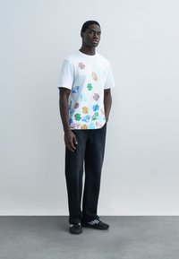 Mann som står mot en enkel vegg iført hvit T-skjorte med fargerike dollartegn og diamanter, svarte bukser og svarte joggesko.