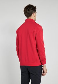 Czerwona bluza z wysokim kołnierzem, wykonana z miękkiego materiału. Posiada ściągacze u mankietów i u dołu oraz gładką fakturę, pokazana z tyłu.