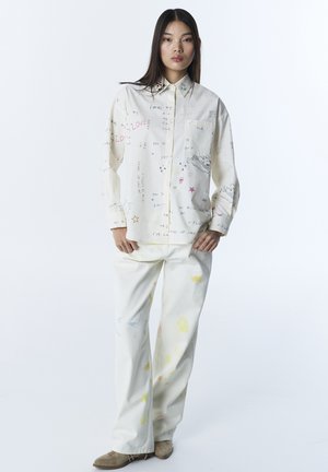 Młoda kobieta stoi, ubrana w luźną białą koszulę z kolorowym, ręcznie napisanym tekstem oraz kremowe spodnie z jasnymi plamami farby, beżowe buty.