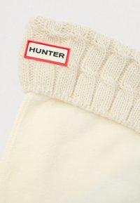 Kremfargede fleece-sokker med en teksturert strikket kremfarget mansjett. Har et rødt og hvitt rektangulært merke merket "HUNTER." Myk og varm design.