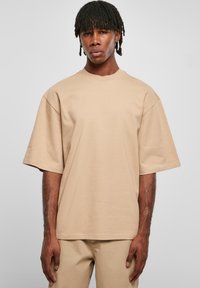 Tricou oversized bej din bumbac; are guler rotund, umeri căzuți și mâneci scurte. Pe partea de jos este drept, cu un design minimalist.
