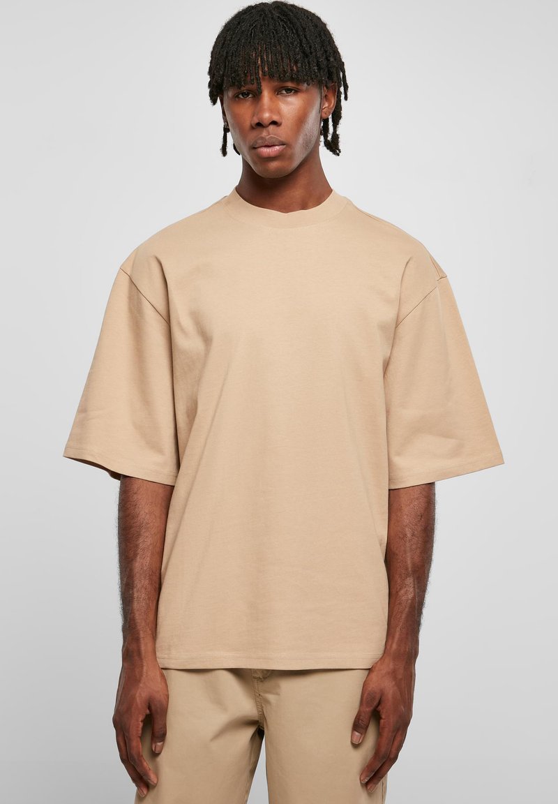 Tricou oversized bej din bumbac; are guler rotund, umeri căzuți și mâneci scurte. Pe partea de jos este drept, cu un design minimalist.