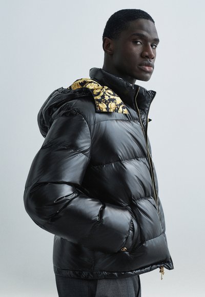 Veste matelassée noire avec une capuche à motifs jaunes et noirs. Elle comprend une fermeture éclair, des poches latérales et une finition brillante.