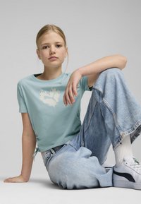 Camiseta corta de color azul claro con un gráfico de llamas en blanco, combinada con pantalones vaqueros sueltos de denim claro. La ropa presenta lazos laterales sutiles y una tela texturizada.