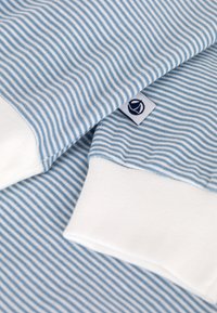 Lichtblauwe en witte gestreepte stof met ribbeltextuur, voorzien van witte manchetten en een klein logo-label in marineblauw aan de onderrand.