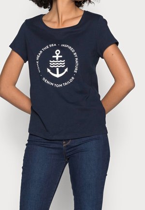 Kvinde iført navyblå T-shirt med hvid anker- og bølgedesign og teksten "Home near the sea, inspired by nature, Denim Tom Tailor", kombineret med mørke jeans.