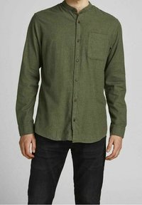 Man som bär en grön långärmad skjorta med knappar och bröstficka samt svarta byxor mot en neutral bakgrund.