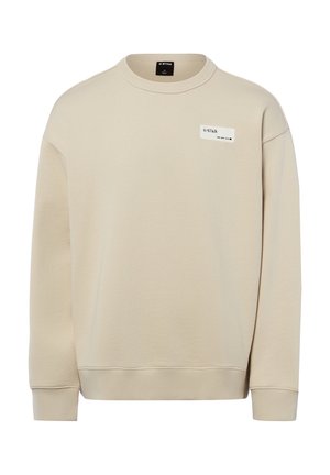 Beżowa bluza z długim rękawem i okrągłym dekoltem, z prążkowanymi ściągaczami przy mankietach i dole oraz małą białą prostokątną naszywką z logo na górnej części piersi.