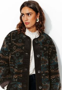 Veste en polaire avec un motif floral texturé en marron foncé, turquoise et beige, dotée d'une finition en velours côtelé noir et de quatre boutons de fermeture.