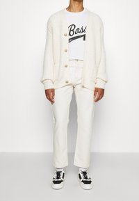 Krämfärgad stickad cardigan med stora knappar, bärs över en vit t-shirt med en svart grafik, ihop med vita raka jeans och svarta sneakers.