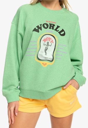 Grøn sweatshirt med teksten "WORLD Surfing Tour" og en sort-hvid surfergrafik, sat sammen med gule shorts med teksten "ROXY."