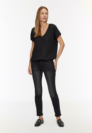 Blouse noire à col en V avec des manches courtes, associée à un jean en denim foncé. Modèle portant des mocassins noirs. Matière simple et lisse avec un design ajusté.