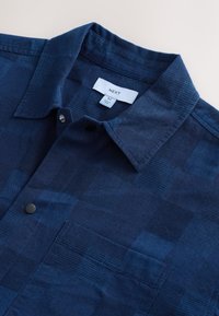 Camicia blu navy con bottoni realizzata in tessuto testurizzato con motivo a quadri. Presenta un colletto appuntito e una tasca singola sul petto. Etichetta del marchio visibile.