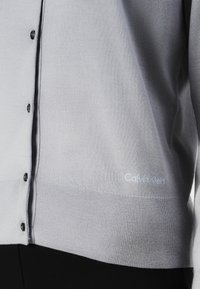 Cardigan cinza tricotado com botões pretos; apresenta uma bainha canelada e o logo "Calvin Klein" em azul claro no canto inferior esquerdo.
