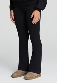 Osoba w czarnych dopasowanych spodniach i beżowych zamszowych butach typu slip-on, stojąca na tle jasnego, jednolitego tła.