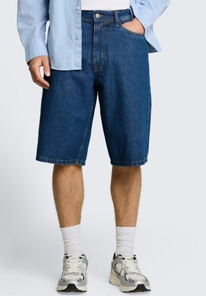 Personne portant un short en jean bleu, des chaussettes blanches, des baskets blanches et une chemise bleu clair à boutons, une main dans la poche.