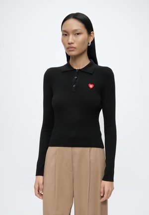 Femme aux cheveux noirs raides portant un polo noir ajusté à manches longues avec un logo cœur rouge et un pantalon beige sur un fond blanc uni.