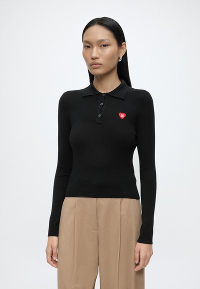 Femme aux cheveux noirs raides portant un polo noir ajusté à manches longues avec un logo cœur rouge et un pantalon beige sur un fond blanc uni.