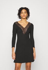 Vestido preto de mangas longas, com decote em V decorado com renda. Parte inferior arredondada com bainha em forma de ondulação. O tecido aparenta ser suave e elástico.