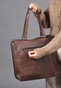 Una persona con un suéter marrón sostiene un bolso tote de cuero marrón oscuro, utilizando la mano derecha para abrir la cremallera del bolsillo frontal exterior, contra un fondo gris.