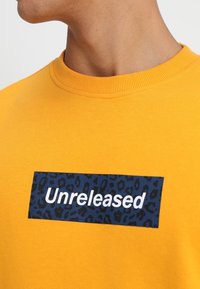 Maglione giallo con scollo rotondo e colletto a costine. Presenta una patch rettangolare blu con stampa leopardata e la parola "Unreleased" in bianco.