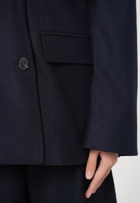 Navyblauer Wollblazer mit einem Ein-Knopf-Verschluss, scharfen Revers und einer Seitentasche, mit glatter Textur und taillierter Passform.