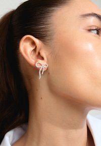 Sudraba auskari, kas veidoti kā loka, ar spīdīgu rhinestone dizainu. Auskariem ir cilpa un karājošas gali.