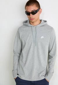 Siva Nike kapuca s prednjim kenguruvim žepom, vezicama i malim belim logotipom na grudima. Model nosi crne naočare za sunce i mornarske pantalone.