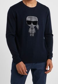 Marinblå stickad tröja med en tecknad karaktär som har solglasögon tryckt i silver. Ribbad mudd och fåll, mjuk textur, figurnära design.