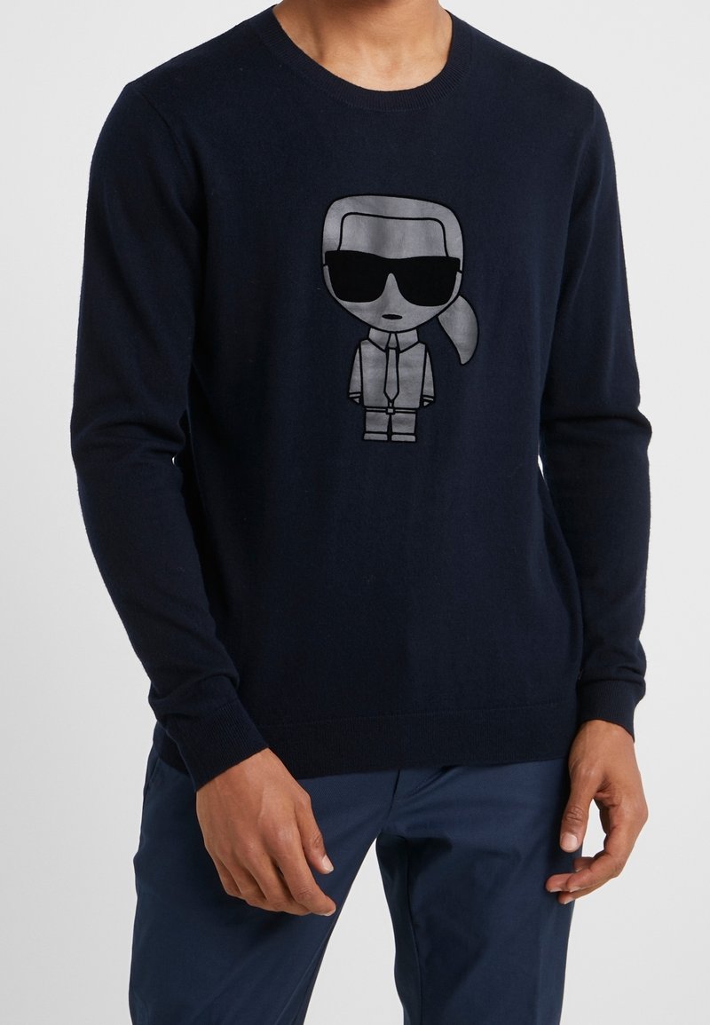 Marinblå stickad tröja med en tecknad karaktär som har solglasögon tryckt i silver. Ribbad mudd och fåll, mjuk textur, figurnära design.