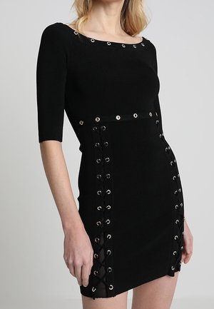 Kobieta w czarnej dopasowanej sukience z metalowymi oczkami i sznurowaniem po bokach oraz przy dekolcie, z rękawami o długości do łokcia.