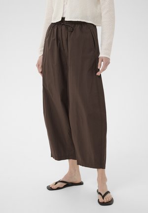 Osoba nosiaca voľné tmavohnedé krátke nohavice, krémový pletený sveter a čierne žabky, stojaca pred obyčajným pozadím.