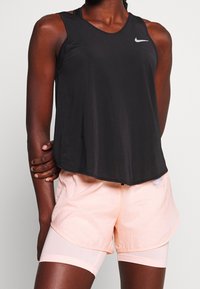 Canotta sportiva nera senza maniche con texture in mesh, con logo Nike argento. Abbinata a pantaloni sportivi rosa chiaro con design a strati.