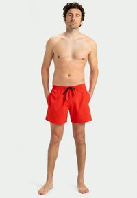 Vyriškis, stovintis basomis, vilkintis ryškiai raudonus maudymosi šortus su juodu stringu, prieš paprastą šviesią foną.