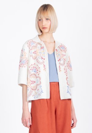 Blondína v bielom krátkom rukáve s gombíkmi a farebnou kvetinovou výšivkou, svetlomodrým topom a hrdzavo sfarbenými nohavicami.