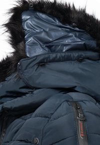 Isolerad marinblå dunjacka med pälsfodrad huva, utrustad med en mörk dragkedja och quiltad textur. Inkluderar en detalj med sidoficka.