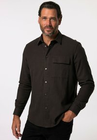 Camicia a maniche lunghe marrone scuro in un tessuto morbido, dotata di tasca frontale e chiusure con bottoni in argento. Colletto strutturato e orlo leggermente curvo.