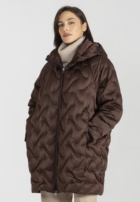 Brązowa ocieplana kurtka z kapturem, z pikowanym wzorem, zapinana na zamek błyskawiczny, z dwiema bocznymi kieszeniami. Nosi ją na lekko beżowym golfie.