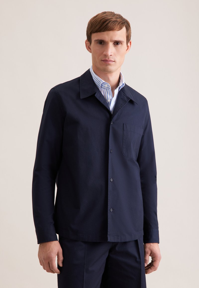 Jeune homme aux cheveux bruns courts portant une veste à boutons bleu marine par-dessus une chemise à rayures bleues claires avec col, debout devant un fond uni.