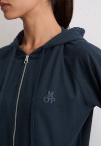 Frau trägt einen dunkelblauen Kapuzenpullover mit Reißverschluss und gestickten "MOP"-Buchstaben auf der Brust, blickt seitlich vor einem hellen Hintergrund.