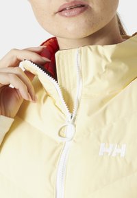 Světlě žlutá zipová bunda s vysokým límcem, texturovaná tkanina a bílý zip. Má červenou podšívku a vyšívané logo.