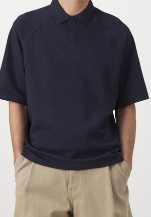 Marineblaues Poloshirt mit kurzen Ärmeln und dreiknöpfiger Knopfleiste, getragen mit beigen Hosen, Hände in den Taschen, keine sichtbaren Logos oder Muster.