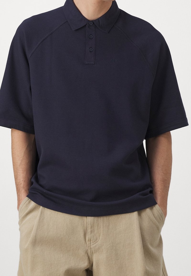 Polo bleu marine à manches courtes avec patte de boutonnage trois boutons, porté avec un pantalon beige, mains dans les poches, sans logos ni motifs visibles.