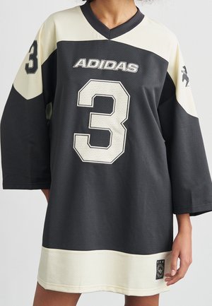 Osoba nosząca oversize czarno-białą sportową koszulkę Adidas z numerem 3 na przodzie i na rękawach, stojąca na jednolitym tle.