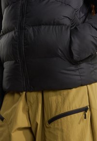 Czarna pikowana kurtka zapinana na zamek, założona na jasnobrązowe spodnie z widoczną czarną kieszenią zapinaną na zamek na prawej udzie.