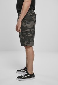Kamouflage cargo shorts in donkergroen en grijs, voorzien van zijzakken en een ontspannen pasvorm. Gecombineerd met zwart-witte lage sneakers.