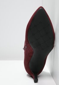 Burgundowe zamszowe szpilki z ostrym noskiem i smukłym obcasem. Podeszwa ma teksturowany czarny wzór. Brak widocznych zdobień.