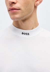 Κοντινό πλάνο ενός άνδρα που φοράει λευκό πουκάμισο με ψηλό γιακά, στο οποίο είναι τυπωμένο το "BOSS" με μαύρα γράμματα στον γιακά, και ένα μικρό κρίκο σκουλαρίκι στο αριστερό αυτί.