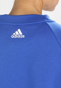 Camisa atlética azul con una textura suave, que presenta un logo blanco de Adidas en la parte posterior cerca del escote. Diseño de cuello redondo.
